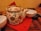 An ornately decorated ceramic teapot with floral and bird designs rests on a red napkin placed on a wooden table. It has a bamboo handle and is surrounded by white bowls, one of which contains a spoon. A plate of cookies sits nearby, adding a cozy touch.
