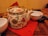 An ornately decorated ceramic teapot with floral and bird designs rests on a red napkin placed on a wooden table. It has a bamboo handle and is surrounded by white bowls, one of which contains a spoon. A plate of cookies sits nearby, adding a cozy touch.