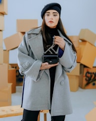 A woman stands confidently holding a small black handbag, wearing a stylish gray coat and a black beret. The background features several stacked cardboard boxes, suggesting a warehouse or moving setting.