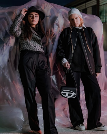 Two individuals pose stylishly against a backdrop of translucent material. One person wears a black hat and a patterned top, while the other sports a white beanie and a dark jacket. The scene is dimly lit with hints of pink and white lighting.