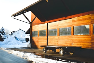 A wooden building with a large, exposed overhang and multiple windows reflecting light. In front of the building, an empty flatbed trailer is parked on a paved surface. Snow covers the surroundings, including a pile near a house visible in the background, suggesting a winter setting. Sunlight casts long shadows and highlights the warm tones of the wood.