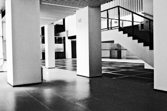 A modern, minimalistic interior space featuring large vertical columns and an open staircase with a black railing. The floor is tiled in a grid pattern, and the ceilings have a linear wooden design.