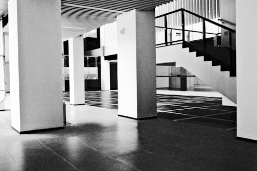 A modern, minimalistic interior space featuring large vertical columns and an open staircase with a black railing. The floor is tiled in a grid pattern, and the ceilings have a linear wooden design.