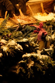 A festive winter scene featuring a decorated food stall. The stall has a garnish of snow-covered fir branches and red berries with a red ribbon. In the background, there are wooden spatulas placed in pots containing various hot dishes. A person wearing dark gloves can be seen serving food.