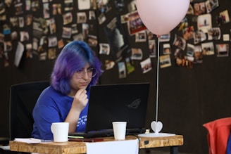 A person with colorful hair, wearing glasses and a blue shirt, is focused on working in front of a laptop. There's a decoration stand with a small heart at the base and a pink balloon tied to it on the desk nearby. The background consists of a black wall covered with numerous pinned photographs and notes.