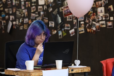 A person with colorful hair, wearing glasses and a blue shirt, is focused on working in front of a laptop. There's a decoration stand with a small heart at the base and a pink balloon tied to it on the desk nearby. The background consists of a black wall covered with numerous pinned photographs and notes.