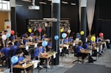 A group of people dressed in blue shirts sit at tables in a modern, open-space office environment. They are focused on their laptops, with paperwork and personal items scattered on desks. Balloons of various colors are tied to the tables, and a large projector hangs from the ceiling. The backdrop displays a wall with the words 'Community is Everything' and photo collages.