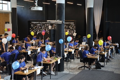 A group of people dressed in blue shirts sit at tables in a modern, open-space office environment. They are focused on their laptops, with paperwork and personal items scattered on desks. Balloons of various colors are tied to the tables, and a large projector hangs from the ceiling. The backdrop displays a wall with the words 'Community is Everything' and photo collages.