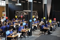 A group of people dressed in blue shirts sit at tables in a modern, open-space office environment. They are focused on their laptops, with paperwork and personal items scattered on desks. Balloons of various colors are tied to the tables, and a large projector hangs from the ceiling. The backdrop displays a wall with the words 'Community is Everything' and photo collages.