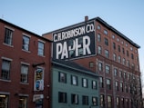 A multi-story brick building with a prominent, large white and black painted advertisement for 'C.H. Robinson Co. Paper' on its side. Nearby, there is a smaller building with a sign for 'Old Port Tavern' hanging from it. The sky is clear, contributing to a peaceful urban scene.