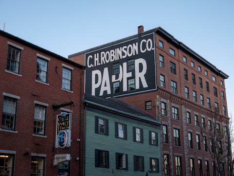 A multi-story brick building with a prominent, large white and black painted advertisement for 'C.H. Robinson Co. Paper' on its side. Nearby, there is a smaller building with a sign for 'Old Port Tavern' hanging from it. The sky is clear, contributing to a peaceful urban scene.