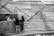 Three children are interacting in an environment constructed with corrugated metal sheets and wooden planks. Two of the children are standing on one side of the metal barrier, while a third child, holding a piece of paper, looks over from the other side. The setting appears to be makeshift, possibly an informal settlement or construction site.