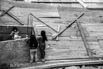Three children are interacting in an environment constructed with corrugated metal sheets and wooden planks. Two of the children are standing on one side of the metal barrier, while a third child, holding a piece of paper, looks over from the other side. The setting appears to be makeshift, possibly an informal settlement or construction site.