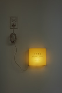 A glowing square light fixture is mounted on a wall, with the words 'Suzuki Shop' printed on it. A cord connects the fixture to an electrical outlet above it. The setting gives a minimalist and modern aesthetic.