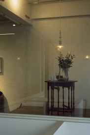 A dimly lit interior space with a wooden table topped with a vase of flowers and a ceramic cup. A suspended pendant light hangs above the table, casting soft illumination. The atmosphere is minimalist with light-colored walls and a calm, serene ambiance.