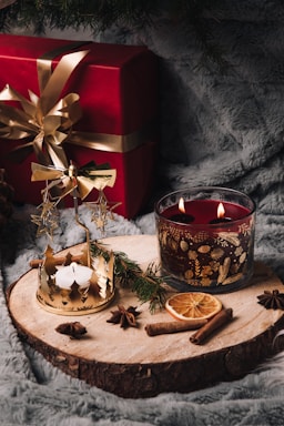 A cozy holiday scene featuring a red gift box with a golden ribbon, a lit candle in a decorative glass holder with pinecone patterns, and a small golden carousel with star motifs. The items are placed on a wooden slab surrounded by festive elements like dried orange slices, cinnamon sticks, star anise, and pine sprigs, all set on a soft, gray plush surface.