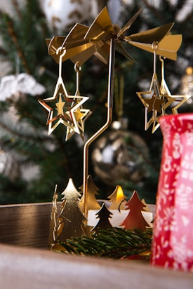 A festive holiday scene featuring a decorative metallic carousel with star-shaped ornaments. A Christmas tree with blurred decorations serves as the background, adding to the festive atmosphere. A red cup with white patterns is partially visible in the foreground.