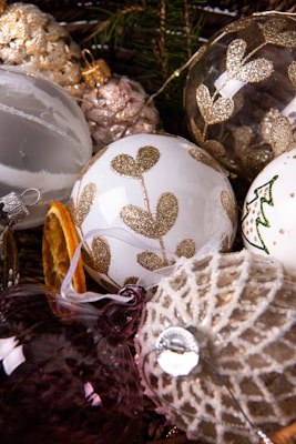 A collection of decorative Christmas ornaments is displayed, featuring white, gold, and silver baubles with glittery patterns and embellishments. Pine cones, a slice of dried orange, and evergreen branches add a festive touch. The ornaments vary in texture, including smooth surfaces and intricate lace-like designs.