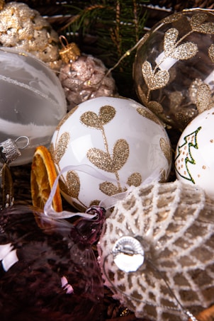 A collection of decorative Christmas ornaments is displayed, featuring white, gold, and silver baubles with glittery patterns and embellishments. Pine cones, a slice of dried orange, and evergreen branches add a festive touch. The ornaments vary in texture, including smooth surfaces and intricate lace-like designs.
