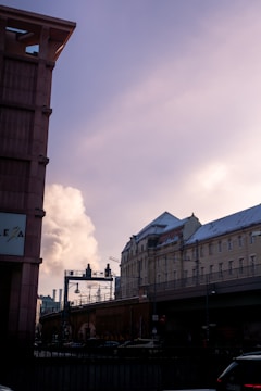 Urban scene featuring a mix of old and modern architecture with buildings and infrastructure elements. A plume of steam or smoke rises in the background against a cloudy sky, while parts of the buildings have snow on them. A road with a bridge passes overhead.