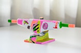A skateboard truck painted with vibrant, abstract patterns including symbols like an 'at' symbol, hashtag, and bold lines. The color scheme includes bright pinks, greens, yellows, and oranges with a glossy finish. The truck is positioned on a white surface with a blurred, neutral background.