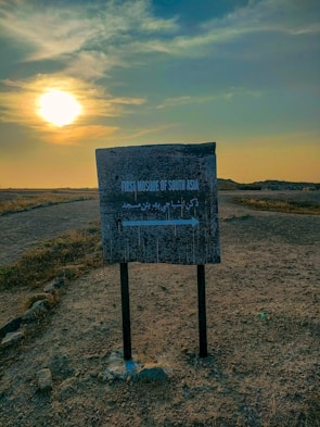A weathered sign stands in a barren, open landscape with a dirt path leading into the distance. The sign reads 'First Mosque of South Asia' in both English and an unknown script, with an arrow pointing to the right. The sun is setting, casting a warm glow over the scene, and the sky is partially clouded.