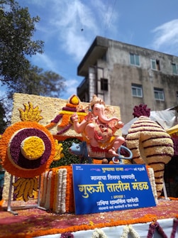 An ornate sculpture of the Hindu deity Ganesha is adorned with colorful floral arrangements. The statue is seated with one hand raised in blessing and surrounded by a vibrant display of marigold and other flowers. A blue sign with yellow text is placed in front of the sculpture. In the background, a building with multiple windows is visible under a clear blue sky.