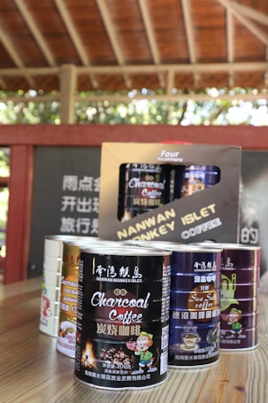 Several cans of flavored coffee, including charcoal and sea salt varieties, are arranged on a wooden table. The packaging features vibrant and colorful designs with cartoon characters. A box in the background displays more coffee cans, and some blurred text appears on a wall.