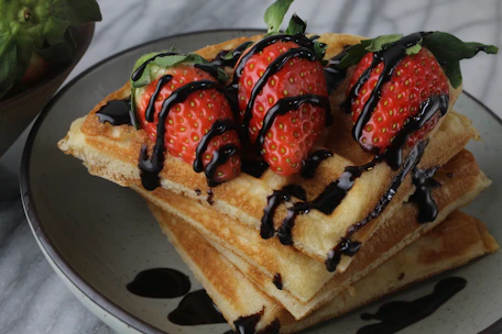 Close-up of a golden waffle topped with fresh strawberries and a drizzle of chocolate syrup on a rustic wooden table