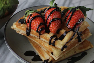 A close-up of a golden waffle topped with fresh strawberries, whipped cream, and a drizzle of chocolate sauce.