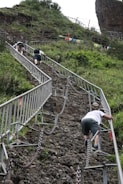 A steep, rocky pathway is being ascended by several individuals using metal railings and chains for support. The path is surrounded by lush greenery and some fencing, and the climbers appear to be focused on their ascent.