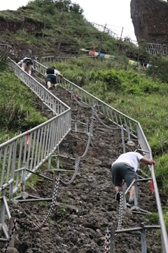 A steep, rocky pathway is being ascended by several individuals using metal railings and chains for support. The path is surrounded by lush greenery and some fencing, and the climbers appear to be focused on their ascent.