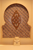 A decorative wall niche featuring an intricate geometric mosaic pattern with vibrant colors. A wooden ornament with an elaborate knot design is mounted centrally. At the base, a small stone sculpture resembling a cat or another small creature sits in front of the mosaic.
