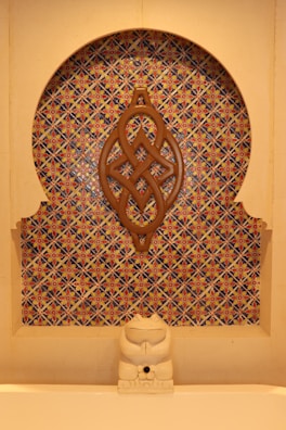 A decorative wall niche featuring an intricate geometric mosaic pattern with vibrant colors. A wooden ornament with an elaborate knot design is mounted centrally. At the base, a small stone sculpture resembling a cat or another small creature sits in front of the mosaic.