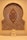 A decorative wall niche featuring an intricate geometric mosaic pattern with vibrant colors. A wooden ornament with an elaborate knot design is mounted centrally. At the base, a small stone sculpture resembling a cat or another small creature sits in front of the mosaic.