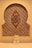 A decorative wall niche featuring an intricate geometric mosaic pattern with vibrant colors. A wooden ornament with an elaborate knot design is mounted centrally. At the base, a small stone sculpture resembling a cat or another small creature sits in front of the mosaic.