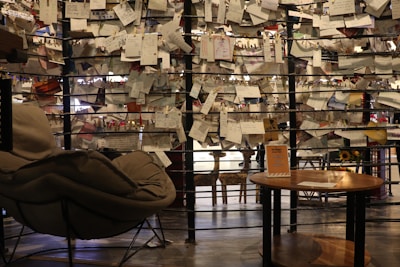 A cozy seating space with a plush chair and a round wooden table in the foreground. A creative backdrop consists of wires holding numerous handwritten notes and papers with clothespins. Sunflowers can be spotted in the background, adding warmth to the environment. Soft, ambient light creates a tranquil and intimate atmosphere.