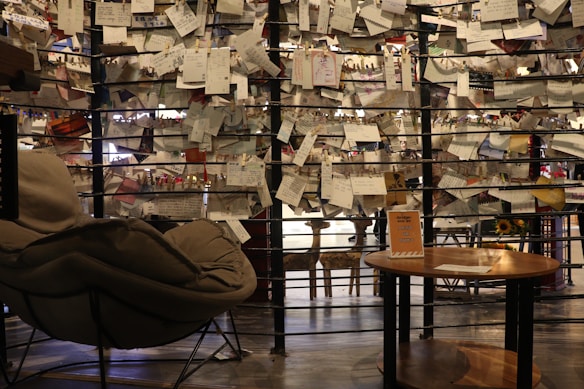 A cozy seating space with a plush chair and a round wooden table in the foreground. A creative backdrop consists of wires holding numerous handwritten notes and papers with clothespins. Sunflowers can be spotted in the background, adding warmth to the environment. Soft, ambient light creates a tranquil and intimate atmosphere.