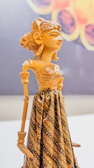 A detailed wooden puppet wearing intricately designed fabric, featuring traditional carvings. The puppet has a carved headdress and facial features that show fine craftsmanship.
