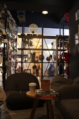 A cozy room interior with dark furniture and a relaxed atmosphere. There are circular armchairs and a small wooden table with a cup and books placed on it. In the background, a shelving unit displays various decorative items including small planters and hanging globes. The lighting is warm, creating a comfortable and inviting environment.