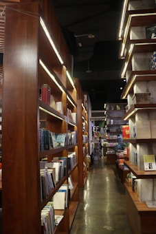 A cozy, dimly lit bookstore corridor lined with tall wooden shelves filled with a variety of books. The polished concrete floor reflects the soft light, creating a warm and inviting atmosphere. Books of different sizes and colors are neatly arranged, inviting exploration and discovery.