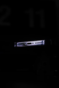 A partially illuminated device edge with various ports and connectors in a dark setting. The focus is on the sleek design of the metallic object, which reflects hints of light, highlighting its contours.