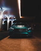 A sleek, metallic car is parked under the warm glow of artificial lights at a modern gas station. The car features a prominent front grille with a recognizable emblem. The station has a minimalist design with several tall, white pillars supporting its canopy.
