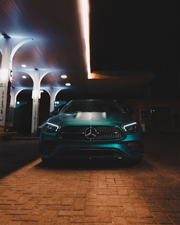A sleek, metallic car is parked under the warm glow of artificial lights at a modern gas station. The car features a prominent front grille with a recognizable emblem. The station has a minimalist design with several tall, white pillars supporting its canopy.