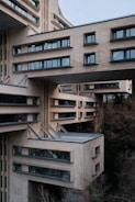An architectural structure with sections of the building extending outward and large, rectangular windows. The design incorporates modern elements with a beige facade, and protruding metal beams. The setting is surrounded by leafless trees and other vegetation, contributing to a somewhat stark and urban atmosphere.