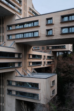 An architectural structure with sections of the building extending outward and large, rectangular windows. The design incorporates modern elements with a beige facade, and protruding metal beams. The setting is surrounded by leafless trees and other vegetation, contributing to a somewhat stark and urban atmosphere.