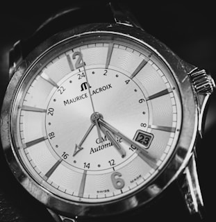 A close-up of an analog wristwatch with bold markers and intricate detailing on the dial. The watch face is elegant, has a date window, and features a combination of numerals and indices. The brand name and other inscriptions are visible, surrounded by a polished metal case.