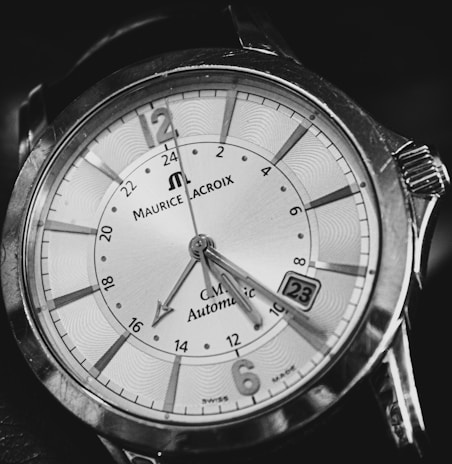 A close-up of an analog wristwatch with bold markers and intricate detailing on the dial. The watch face is elegant, has a date window, and features a combination of numerals and indices. The brand name and other inscriptions are visible, surrounded by a polished metal case.