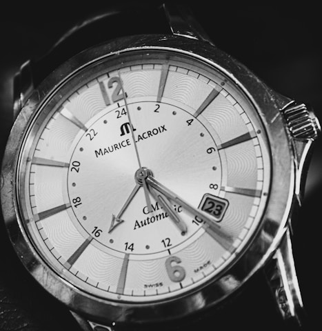 A close-up of an analog wristwatch with bold markers and intricate detailing on the dial. The watch face is elegant, has a date window, and features a combination of numerals and indices. The brand name and other inscriptions are visible, surrounded by a polished metal case.
