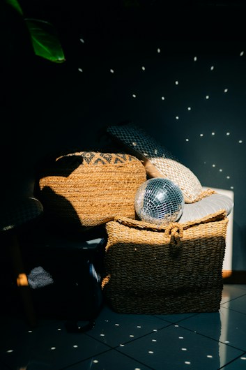 A stylish room corner features a combination of woven baskets and cushions, with sunlight filtering through creating a play of light and shadow. A disco ball adds a reflective touch, casting specks of light across the surface, creating a dynamic and lively pattern.